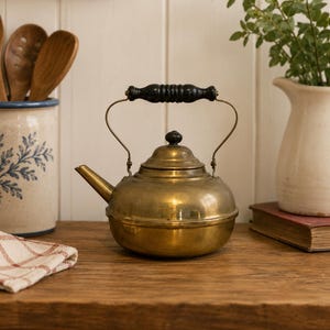 May include: A brass tea kettle with a rounded body, a short spout, and a black ribbed handle. The kettle features a matching black knob on the lid and a metallic finish. The design includes a curved metal frame supporting the handle above the lid.