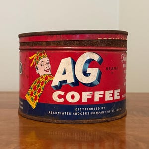 May include: Vintage AG Coffee tin with a red background and white and blue lettering. The tin features a smiling cartoon character wearing a hat and scarf. The text "AG COFFEE" is prominently displayed.