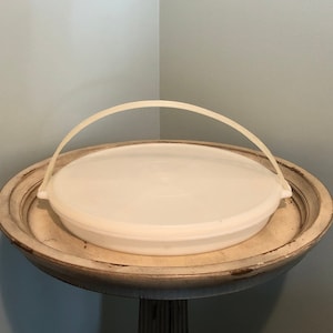 May include: White plastic serving tray with a handle. The tray is round and has a smooth surface. It is sitting on a wooden pedestal.