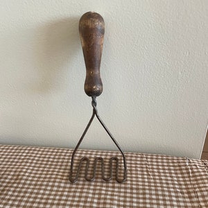 May include: A vintage potato masher with a wooden handle and a wire mashing head. The masher is on a brown and white checkered tablecloth.