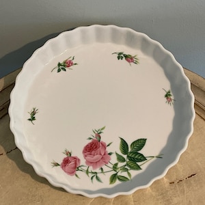 May include: A white ceramic pie plate with a scalloped edge. The plate is decorated with pink roses and green leaves. The floral design is arranged around the center of the plate. The plate is a round shape.