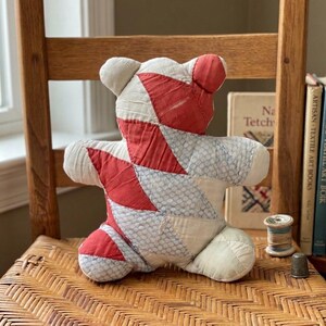 May include: A stuffed bear toy constructed from quilted fabric patches in a diamond pattern. The bear features alternating sections of solid red fabric and light blue fabric with a white grid pattern, with cream-colored fabric on the limbs.