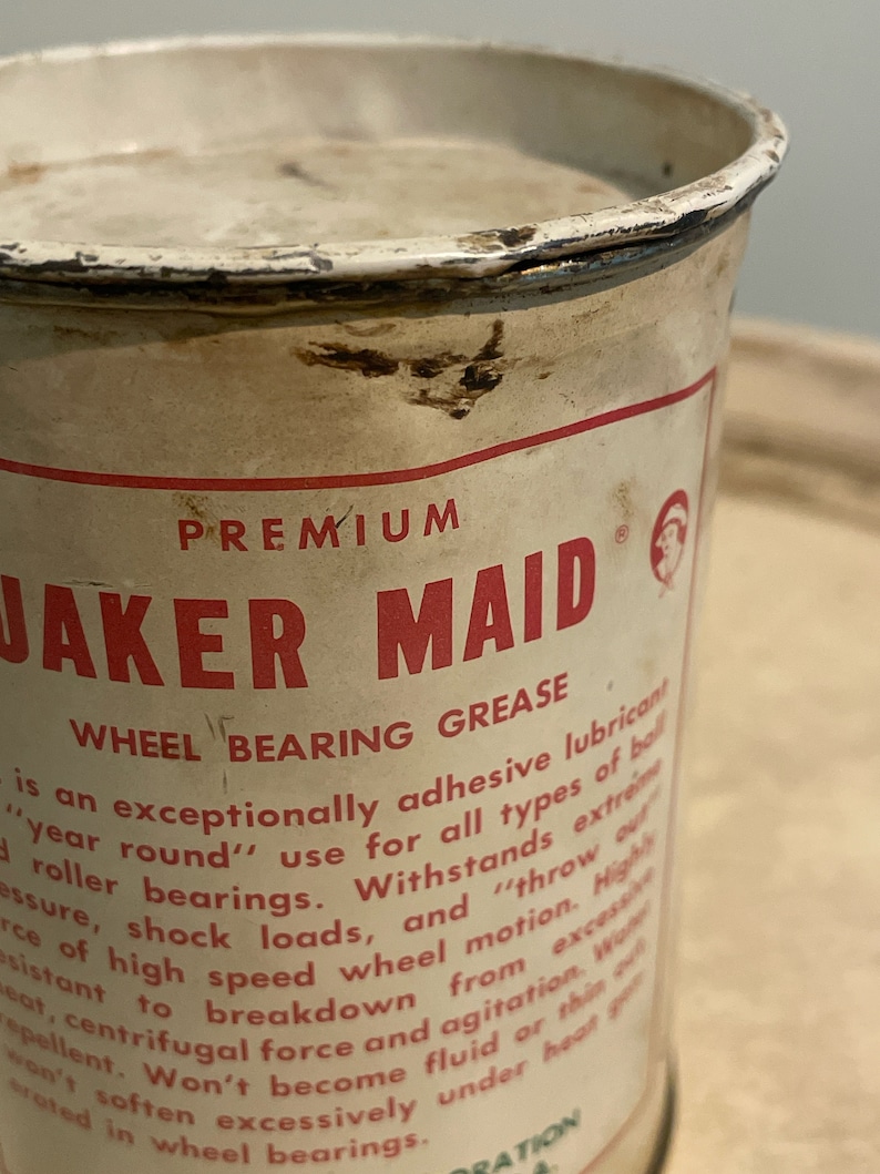 May include: A white metal can with red lettering that reads "PREMIUM JAKER MAID WHEEL BEARING GREASE". The can has a red and white logo with a stylized image of a person. The text on the can describes the product as an "exceptionally adhesive lubricant" for "all types of ball roller bearings".
