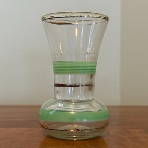 May include: A clear glass vase with a flared rim and a rounded base. The vase features horizontal bands of light green paint and thin, dark brown metallic-toned stripes encircling the neck and the lower body, creating a simple decorative pattern.