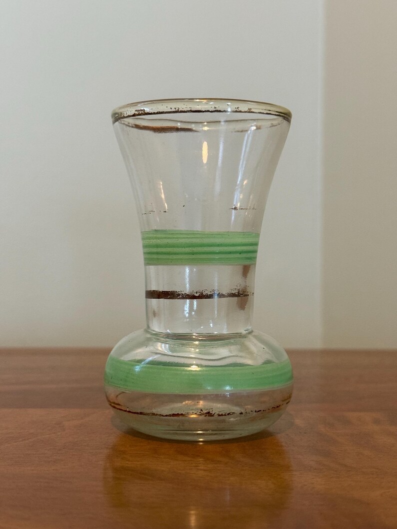 May include: A clear glass vase with a flared rim and a rounded base. The vase features horizontal bands of light green paint and thin, dark brown metallic-toned stripes encircling the neck and the lower body, creating a simple decorative pattern.