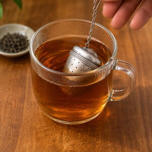 May include: A metallic tea infuser with a perforated body and a small attached chain, submerged in a clear glass mug filled with amber-colored tea. The infuser features a rounded top and a cylindrical base, designed for steeping loose leaf tea.