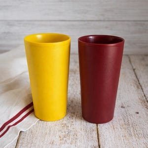 May include: Two tall, cylindrical drinking tumblers standing side by side. One tumbler is a bright, solid yellow color, while the other is a deep, solid maroon red. Both cups feature a smooth, matte finish with a simple, tapered design.