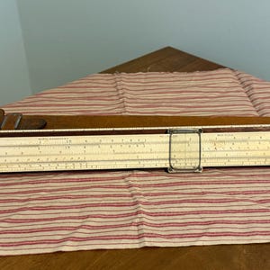 May include: A vintage slide rule with a wooden frame and a cream-colored scale. The slide rule has multiple scales and markings, with a metal frame and a brown leather case. The slide rule is a tool for mathematical calculations.