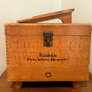 May include: A vintage wooden Ronson Roto-Shine Magnetic shoe shine box with a wooden shoe rest on top. The box is rectangular with a metal clasp and a small handle. The text "Ronson Roto-Shine Magnetic" is printed on the front.