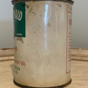 May include: A vintage metal can with a green and white label that says "Maid" and "Guaranteed". The can is labeled "Quik-Drying" and has a red label with text about the product's properties. The can is rusty and has a white background.