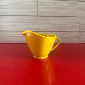 May include: A bright yellow creamer pitcher with a handle and a spout. The pitcher is made of a solid, smooth material and sits on a red surface. The background is a light gray wooden panel.