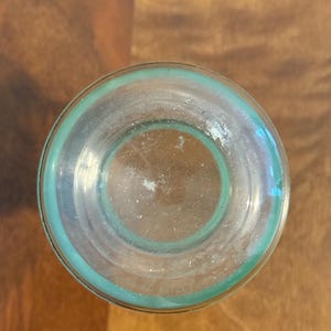 May include: A clear glass vessel viewed from directly above, featuring a circular base with a distinct teal-coloured rim. The glass is transparent, revealing a smooth, rounded bottom surface with subtle light reflections and a clean, simple design.