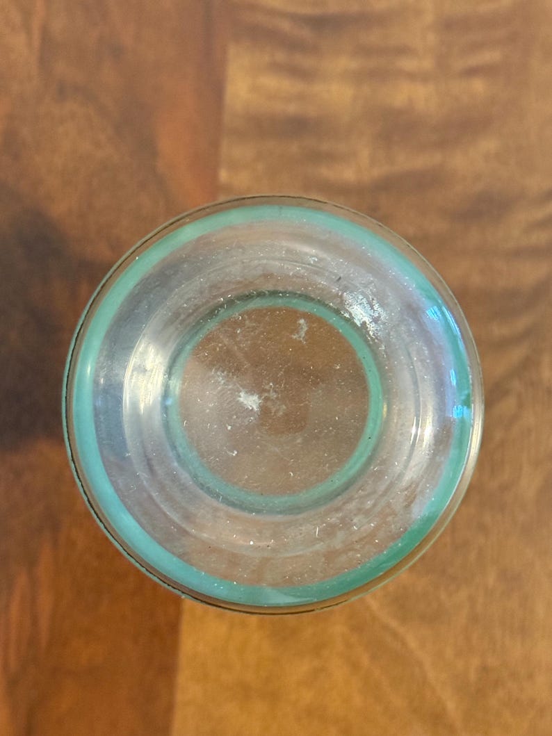 May include: A clear glass vessel viewed from directly above, featuring a circular base with a distinct teal-coloured rim. The glass is transparent, revealing a smooth, rounded bottom surface with subtle light reflections and a clean, simple design.