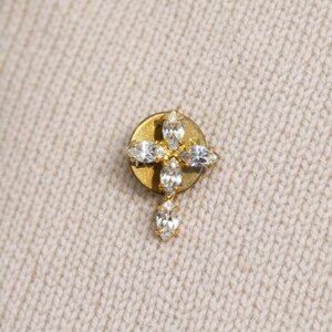 May include: A gold-toned lapel pin featuring a cross design composed of five marquise-cut clear crystals. The cross is mounted on a circular gold-toned backing, creating a structured piece of jewelry with a reflective finish and clear stones.