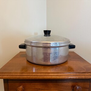 May include: A round, silver-colored metal cooking pot with a matching lid. The pot features two black plastic side handles and a black plastic knob on the center of the lid. The exterior of the pot has three horizontal embossed lines.