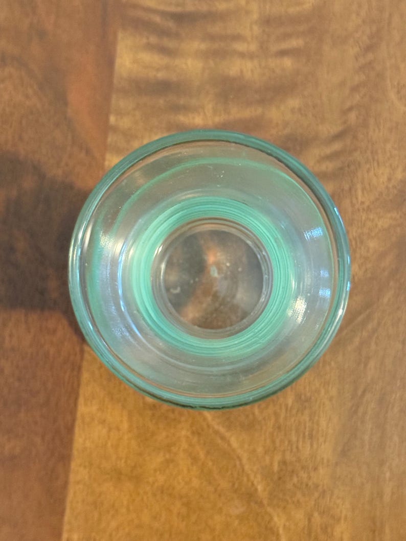 May include: A top-down view of a clear, light green glass vessel with a circular opening. The glass features a thick, rounded rim and a smooth, transparent body that tapers slightly towards the base, showcasing a simple, functional design.