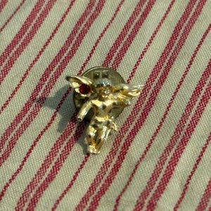 May include: A gold-toned cherub pin with outstretched wings and a red gemstone. The pin is set against a cream-colored fabric with red stripes.