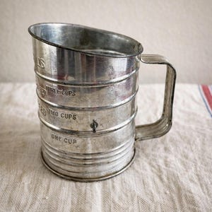 May include: A vintage silver-toned metal flour sifter with a side handle. The exterior features embossed measurement markings for one cup, two cups, and three cups, along with the word SIFTER visible near the top rim of the cylindrical body.