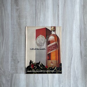 May include: A vintage print advertisement for Johnnie Walker Red Label Scotch whisky. The ad features a bottle of Johnnie Walker Red Label Scotch whisky with a red and gold label. The ad also features the text "Gift of the month." and "Johnnie Walker Red, world's best selling Scotch."