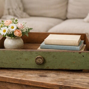 May include: A rectangular wooden tray with a distressed sage green finish and a single centred round knob. Inside the tray sits a small off-white ceramic vase filled with white daisies and peach roses, alongside a stack of two hardcover books.