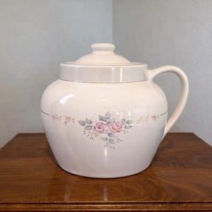 May include: A cream-colored ceramic canister with a matching lid and a single side handle. The body features a delicate floral pattern of pink roses and green leaves centered on the front, with a thin horizontal line encircling the vessel.