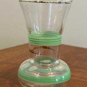 May include: A clear glass vase featuring a bulbous base and a flared rim, decorated with horizontal light green bands and thin brown painted accents around the neck and base. The glass is transparent with a smooth, polished finish throughout.
