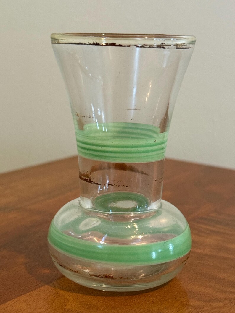 May include: A clear glass vase featuring a bulbous base and a flared rim, decorated with horizontal light green bands and thin brown painted accents around the neck and base. The glass is transparent with a smooth, polished finish throughout.