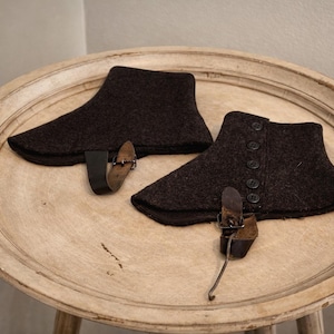 May include: A pair of dark brown felted wool spats featuring a row of five matching buttons along the side and a black leather strap with a metal buckle attached to the bottom edge for securing the garment around the footwear.