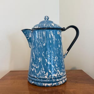 May include: A white ceramic mug with a black handle and the text "But First, Coffee" in a bold, black font. The mug is set against a blurred background. The mug appears to be approximately 10 cm tall.