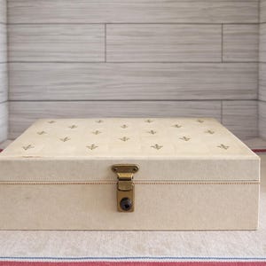 May include: A rectangular cream-colored storage box with a tufted top featuring small gold-toned anchor emblems. The box includes a central brass-colored metal latch mechanism on the front panel for securing the lid, with visible stitching details.