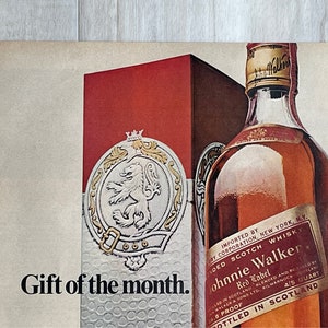 May include: A red and gold box with a lion crest and the words "Gift of the month."  A bottle of Johnnie Walker Red Label Scotch Whisky is in the background. The bottle is labeled "Imported by Cory Corporation, New York, NY. Blended Scotch Whisky. Johnnie Walker Red Label. Bottled in Scotland 4/5 Quart 86 Proof."