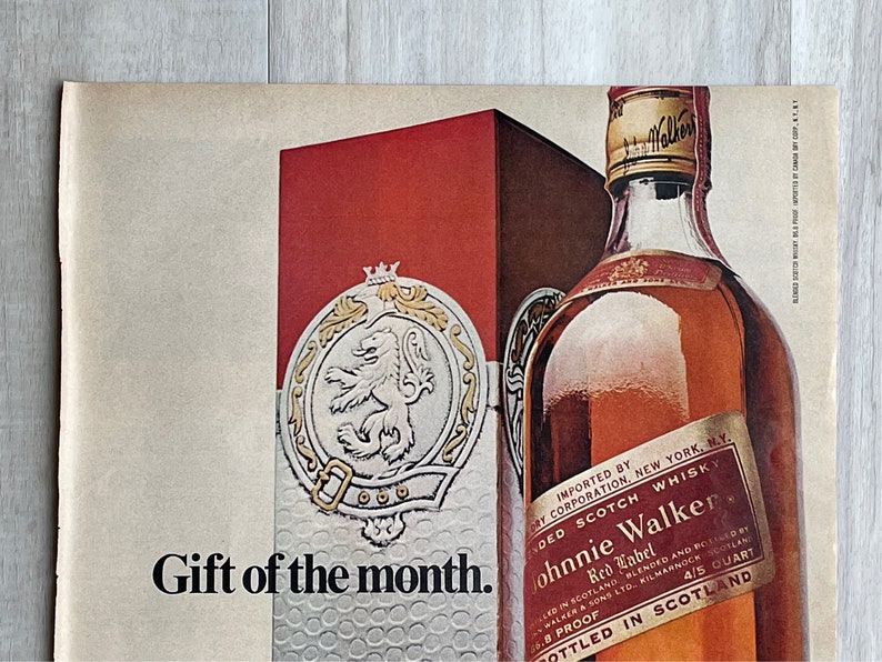 May include: A red and gold box with a lion crest and the words "Gift of the month."  A bottle of Johnnie Walker Red Label Scotch Whisky is in the background. The bottle is labeled "Imported by Cory Corporation, New York, NY. Blended Scotch Whisky. Johnnie Walker Red Label. Bottled in Scotland 4/5 Quart 86 Proof."