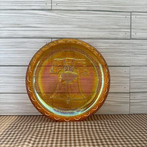 May include: An iridescent, round plate with a detailed image of the Liberty Bell. The plate has a decorative, scalloped edge and text that reads "Liberty Bell Philadelphia". The plate's colors shift from gold to pink.