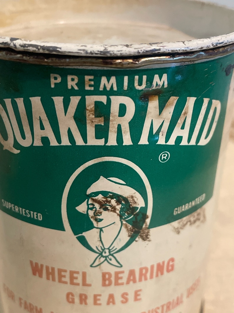 May include: A vintage tin can of Quaker Maid Premium Wheel Bearing Grease. The can is green and white with a red and white logo of a woman wearing a white hat and a green and white sailor collar. The text "SUPER TESTED" and "GUARANTEED" are printed on the can.