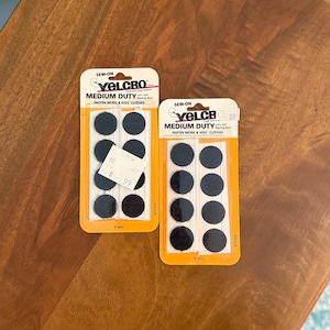 May include: Two packages of black sew-on Velcro brand medium duty hook and loop fasteners. Each package contains four sets of circular fasteners on a white backing card with orange borders. The text reads SEW-ON VELCRO MEDIUM DUTY.