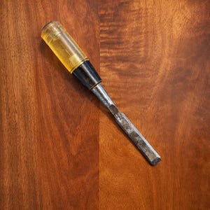 May include: A wood chisel featuring a translucent yellow amber-colored handle, a black ferrule, and a long, flat, rectangular metal blade with a beveled cutting edge. The tool shows signs of wear and age on the metal surface and handle.