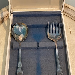May include: A silver serving set in a white box with a blue velvet lining. The set includes a serving spoon and a serving fork.