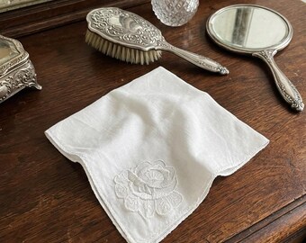 May include: A square white handkerchief featuring a delicate tone-on-tone embroidered rose design in the corner. The fabric is plain white with a subtle texture, and the floral embroidery is stitched in matching white thread, creating a clean look.