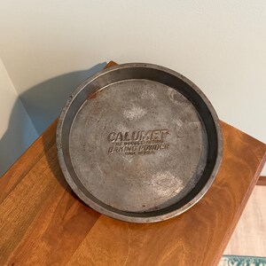 May include: A round, silver-colored baking pan with the text "CALUMET THE DOUBLE-ACTING BAKING POWDER MADE IN USA" embossed on the bottom. The pan is made of metal and shows signs of wear.