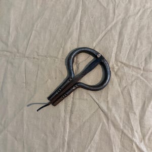May include: A dark metal jaw harp, also known as a mouth harp, lies on a tan fabric surface. The instrument has a curved, U-shaped frame with a thin, flexible metal tongue extending from the center. The harp's design is simple and functional.