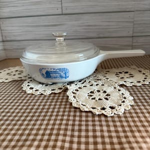 May include: White casserole dish with a clear glass lid and a long handle. The dish features a blue and white illustration of a house and trees. The dish is sitting on a brown and white checkered tablecloth with a cream-colored lace doily.
