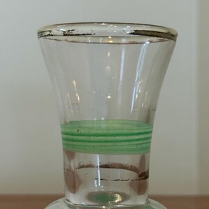 May include: A clear glass vase featuring a flared rim with a worn metallic gold-tone trim. The neck of the vase is decorated with a series of horizontal light green stripes, while the base section also displays a wide light green band.