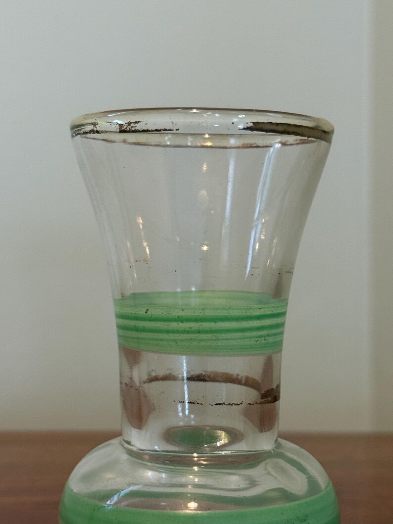 May include: A clear glass vase featuring a flared rim with a worn metallic gold-tone trim. The neck of the vase is decorated with a series of horizontal light green stripes, while the base section also displays a wide light green band.