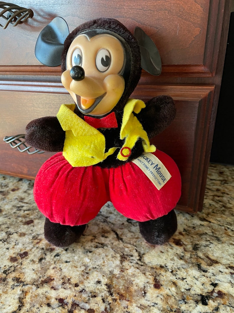 May include: A plush Mickey Mouse toy with a red and yellow outfit. The toy has a black nose, large black eyes, and a red bow tie. The tag on the toy reads 'Mickey Mouse' and 'Walt Disney Productions.'