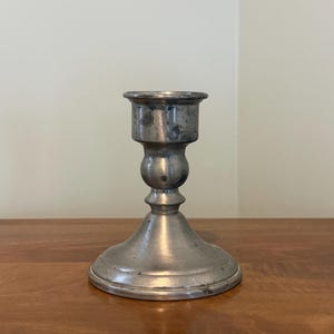 May include: A vintage silver candlestick holder with a wide, circular base and a bulbous midsection. The top has a small, shallow cup to hold a candle. The metal shows signs of age and wear, adding to its antique charm.