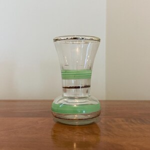 May include: A small clear glass vase featuring a bulbous base and a flared rim. The vase is decorated with horizontal light green painted stripes and thin metallic gold bands around the neck, the midsection, and the base of the glass vessel.