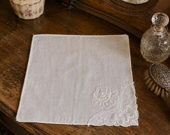 May include: A square white handkerchief featuring delicate white floral embroidery in one corner. The fabric has a visible weave texture and a finished stitched edge. The embroidery design includes a central flower surrounded by scrolling leaf patterns.
