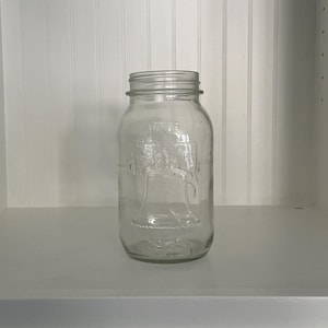 May include: A clear glass jar with a wide mouth and a lid. The jar has a textured surface with the words "1776" and "Liberty Bell" embossed on the side.