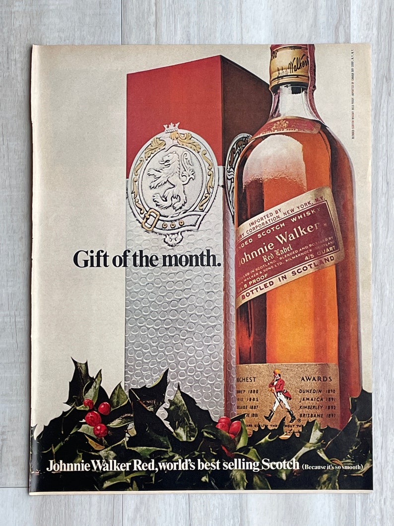 May include: A vintage advertisement for Johnnie Walker Red Label Scotch whisky. The ad features a bottle of the whisky, a red box, and a festive holly sprig. The text reads "Gift of the month." "Imported by Dry Corporation New York, NY." "Blended Scotch Whisky." "Johnnie Walker Red Label." "4/5 Quart." "Blended and Bottled in Scotland." "86 Proof." "Chest 1880, 1883, 1885, 1887." "Awards Dunedin 1890, Jamaica 1891, Kimberley 1892, Brisbane 1897." "Johnnie Walker Red, world's best selling Scotch (Because it's so smooth)"