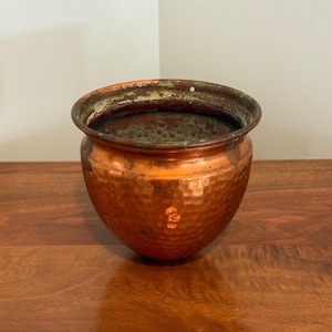 May include: A round, hammered copper pot with a wide, open top and a slightly flared rim. The exterior features a textured, dimpled surface with a warm metallic finish, while the interior shows signs of natural oxidation and patina.